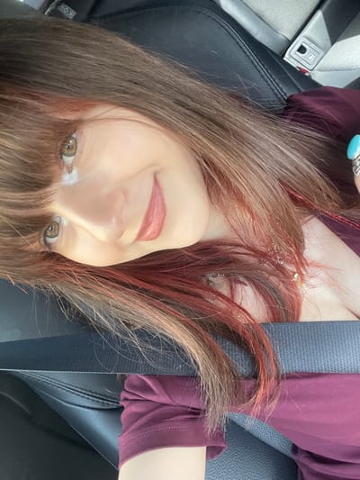 A single female individual is depicted in a close-up, upward-angled self-portrait within a vehicle in Kfar Saba, Israel. The subject, positioned with her head tilted to the left, wears a dark maroon long-sleeved shirt and has long brown hair with prominent reddish-auburn highlights, styled with bangs. Her eyes are light-colored, and she displays a closed-lip smile. A silver-toned necklace with small charms is visible around her neck, and a silver-toned ring with a large turquoise-colored stone is present on her left ring finger, partially visible at the upper right. She is secured by a dark-colored seatbelt fastened across her chest and lap. The immediate environment consists of a black vehicle seat, likely leather or synthetic, with visible stitching and a seatbelt buckle mechanism.