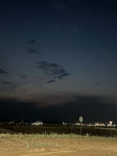 The image depicts a nighttime scene on the outskirts of Bozshakol, Kazakhstan. The sky is dark with scattered clouds, and a faint light emanates from a distant settlement or event, visible as a line of lights along the horizon. A single bright star or planet is visible in the sky.
In the foreground, a dirt path runs along a grassy embankment. Several vehicles are parked along the side of the path, including a white SUV and a few other dark-colored cars. Some of the cars appear to have lights on, possibly for illumination or as part of an event. Two road signs are visible on poles; one is a circular sign with a red border and a diagonal line, indicating a prohibition, and another is a triangular sign with a red border and a white center, typically used for warning.
The overall impression is one of a rural or semi-rural setting at night, possibly near a gathering or a temporary event, given the parked vehicles and distant lights. The time of day is clearly night, and the weather appears calm. There is no visible text in the image.