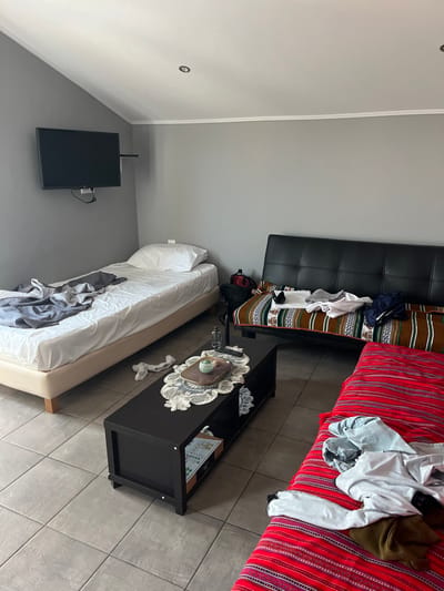 The image shows a living room in a small house or apartment located at 40.627 latitude and 24.575 longitude in Limenaria, Greece. The room features gray walls, a tiled floor, and a sloped ceiling. There is a single bed in the left corner of the room with a gray blanket on top and a white pillow. To the right of the bed is a black coffee table with a white doily and a few items on top. Next to the coffee table is a black leather couch with a colorful blanket and a few items of clothing on it. In the far right corner of the image is a couch covered in a red and white patterned fabric with several items of clothing on it. A flat screen TV is mounted on the wall behind the bed. The room appears to be in a state of disarray.