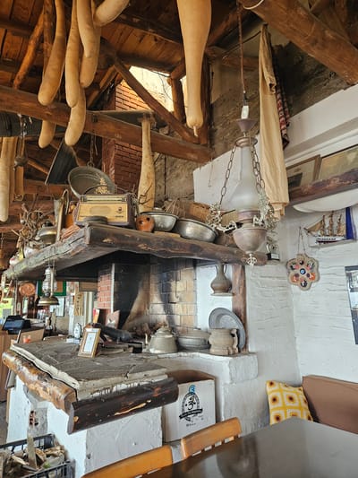 This image depicts the rustic interior of a establishment in Tire, Turkey, characterized by traditional architecture and an abundance of decorative and functional objects.
The scene is dominated by a large, brick fireplace structure with a prominent, dark-stained wooden mantelpiece and a lower cooking surface. The ceiling features exposed, rough-hewn wooden beams from which numerous elongated, light-colored, possibly dried gourds or plant husks, are suspended. A vintage-style chandelier with a frosted glass shade and chains hangs centrally, with a wall-mounted light switch visible nearby.
On the mantelpiece, a vintage radio with a light-colored grille and dark casing is prominently displayed, surrounded by various metal pots, a textured ceramic vessel, and decorative dried branches. Below the mantel, the fireplace opening shows sooted bricks, and to the left, a large, flat stone slab serves as a cooking surface or hearth. On this slab, a metal kettle, a framed certificate, and other miscellaneous items are arranged. A wooden log accent runs along the front of this lower structure.
The walls are predominantly white-washed, contrasting with exposed brick sections of the fireplace and the dark wood beams. On the right wall, a model wooden sailing ship is displayed on a small shelf, and below it, a circular, multi-colored decorative wall hanging featuring several "evil eye" motifs is suspended. The left side of the room shows a glimpse of a modern electronic register system.
In the foreground, a dark, reflective wooden table is visible, flanked by wooden chairs. A seating area with a plain brown cushion and a yellow crocheted cushion adorned with sunflower patterns is against the far right wall. A cardboard box labeled "Tequila Tres Magueyes BLANCO" rests on the floor near the table, and a black crate filled with wood scraps and possibly tools is also visible on the floor to the left. Sunlight enters the space from an unseen opening towards the upper left, highlighting the rustic textures and objects.