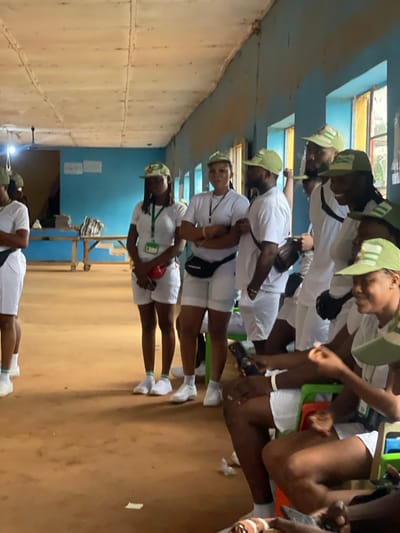 In Ise-Ekiti, Nigeria, an indoor scene captures a group of individuals in white uniforms and green caps, suggestive of a national service or youth program. The room features light blue walls, a light-colored, paneled ceiling, and natural light filtering through windows. The floor is a dusty, reddish-brown earth.
Several young adults are present, mostly standing, with some seated. They appear to be engaged in conversation or waiting. The individuals are dressed in white t-shirts, white shorts, and matching green baseball caps. Some wear lanyards with identification cards. One woman in the foreground is holding a red pouch around her waist. In the background, a wooden table with some items on it is visible. The overall atmosphere suggests a gathering or orientation session.