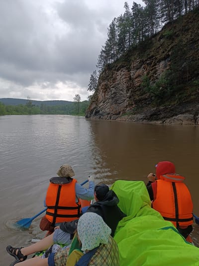 An overcast day, a raft with four people drifts on a muddy river. Two people in orange life vests are visible from the back, facing forward in the raft. One person is paddling with a blue oar. The raft is covered with a bright green tarp. The riverbank on the right is a steep, rocky cliff covered with moss and trees. The cliff face is dark and textured with visible layers of rock. On the left side of the river, a dense forest stretches to the horizon, leading to rolling hills under a cloudy sky. The water is a murky brown, reflecting the overcast sky. The scene appears to be in a natural, undeveloped area, possibly the countryside near Idrisovo, Russia, given the natural landscape and vegetation.