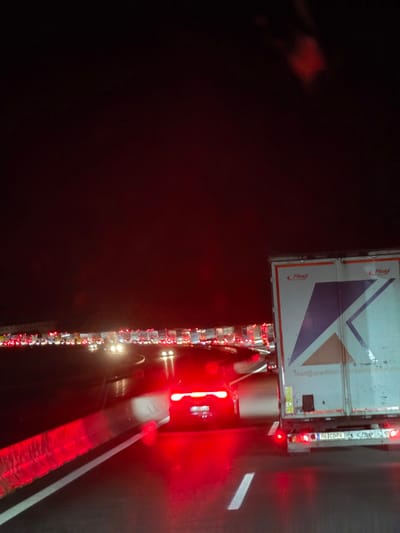 The image depicts a nighttime scene of heavy traffic congestion on a highway. In the foreground, a red car with its brake lights illuminated is stopped in traffic. To the right, the rear of a white truck with a large "K" graphic and the logo "Fliegl" is visible. The truck displays text that appears to be an email address: "mail@spedition-..." and a license plate that reads "BLCD-74". In the background, a long line of trucks and cars are stationary, with their red taillights creating a glowing effect against the dark, possibly wet, road surface. The sky is dark, indicating it is night. There is no indication of people's presence or specific interactions beyond the stopped vehicles. The location is identified as Uettingen, Germany, likely on a highway.