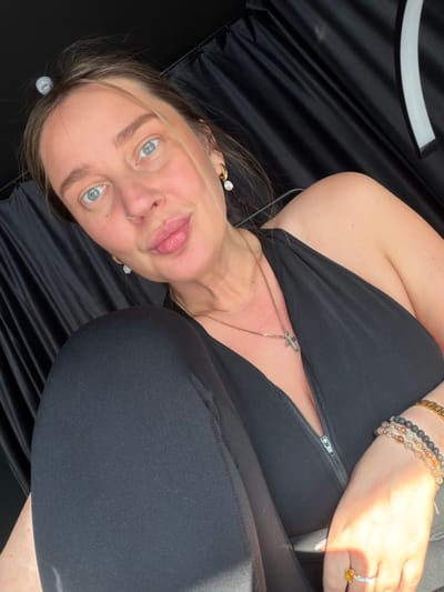 A woman with light blue eyes and light brown hair is pictured laying down on her side, with her face towards the camera. She is wearing a black top, black leggings, and several bracelets on her wrist. She is also wearing jewelry on her neck, ears, and nose. The background appears to be dark and flowing fabric, with a white curved object visible at the top left. The lighting suggests it is daytime, with sunlight illuminating the woman's arm and hand. The woman's expression is neutral, and her gaze is directed at the camera. There are no visible text or specific location cues in the image.
