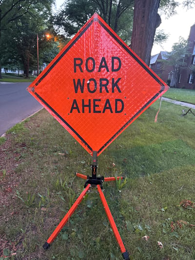 The image shows a diamond-shaped orange road work sign with black lettering that reads "ROAD WORK AHEAD". The sign is mounted on a black and orange tripod stand and is positioned on a grassy area next to a paved road. In the background, there are trees, houses, and a street light. The scene appears to be in a residential neighborhood during the evening or early morning, as indicated by the soft lighting and the lit street light. The sign is reflective and appears to be made of metal or a similar durable material. The tripod stand is made of metal and has orange legs. The grassy area is unkempt and contains some fallen leaves and weeds. The road is a typical asphalt street. The houses in the background appear to be older, possibly from the mid-20th century. The overall impression is that of a temporary traffic control measure in a suburban setting. There are no people or vehicles visible in the image. The photograph was likely taken in Shaker Heights, Ohio, United States of America, given the stylistic and architectural cues of the houses and the presence of mature trees, which are characteristic of the area.