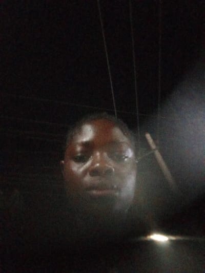 A dimly lit, blurry selfie of a person with dark skin. The individual is centered in the frame, and only their head and shoulders are visible. In the background, there are dark vertical lines, possibly utility poles or wires. A bright light source is visible in the lower right corner. The location, Mbribit, Nigeria, is not discernible from the visual information.