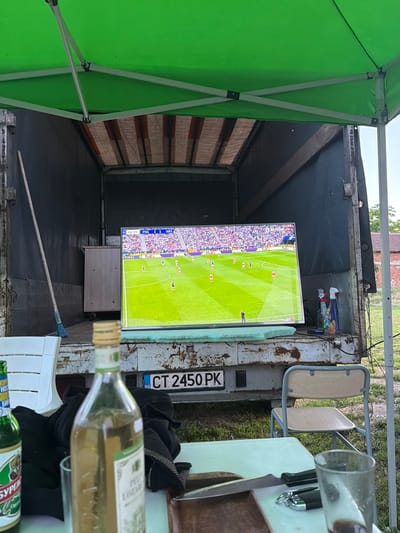 The image shows an outdoor setup within the open back of a truck, seemingly in Malak dol, Bulgaria (based on the license plate "CT 2450 PK"). A television screen is prominently displayed, showing a soccer match (PSG vs. ARS with a score of 1-1 at 86:58). The truck's interior is utilitarian, appearing worn and somewhat dirty, with items stored in the upper section. A broom lies on the floor.
Outside the truck, a green canopy provides shade. A folding chair is visible near the truck bed. A white table partially obscures a bottle of white wine, a beer bottle, a glass, cutlery, and a cutting board. Black cloth is draped over or beside these objects. The surrounding environment includes grass and possibly a building in the background.
