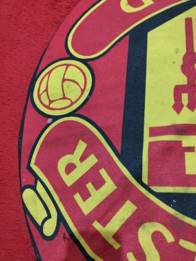 This image presents an angled, close-up forensic view of a section of a Manchester United football club logo, photographed in Lumajang, Indonesia. The logo is printed on what appears to be a red, textured fabric item. The design is predominantly red and yellow with black outlining. Visible elements include the upper left portion of a yellow football graphic contained within a red circular field. Below this, the yellow letters "STER" (from "MANCHESTER") are discernible on a red, scroll-like banner section. To the right, a segment of the club's yellow devil/pitchfork emblem is visible against a red background, bordered on its left by a prominent vertical black line. Minor surface wear, including subtle smudges and faint specks, is present across the yellow and red printed areas of the logo. No individuals or active interactions are depicted.