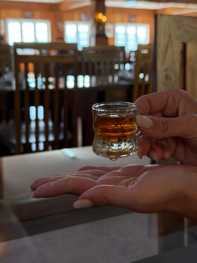 A small, clear glass vessel, containing an amber-colored liquid and a dark, slender object, is held by the thumb and index finger of a hand with light-to-medium skin tone and manicured, pale-colored fingernails. A single drop of liquid is suspended from the bottom of the glass. Directly beneath the glass, the open palm of another hand, displaying similar skin tone and nail characteristics, is positioned. The blurred background depicts an indoor establishment, likely a restaurant or cafe, with multiple wooden chairs, tables, and visible wooden architectural elements. Bright natural light enters through several rectangular windows. The scene is located in an interior space within the city of Ramboda, Sri Lanka.