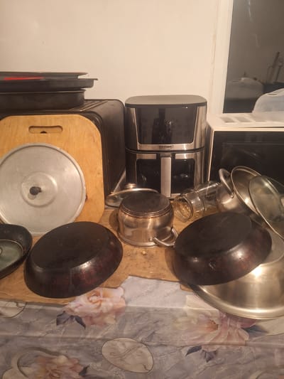 The image displays a collection of kitchenware, primarily pots and pans, arranged on a wooden surface. A coffee maker with the brand name "ТЕХНОМИР" is visible on the left. In the background, a microwave oven and a patterned tablecloth with floral designs can be seen. The setting appears to be a kitchen in a new building ("новостройка") in Kyrgyzstan. The lighting suggests it might be indoors, possibly in the evening. There are no people present in the image, and the overall mood is neutral, focusing on the objects.
