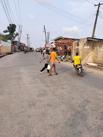 The image depicts a bustling street scene in Ojodu, Nigeria, under a partly cloudy sky. The asphalt road occupies the foreground and midground.

In the foreground, three individuals are present. Two females are walking across the street from the left side towards the right. The lead female wears an orange t-shirt, light blue denim shorts, and dark sandals, with her hair styled in braids. The second female, walking slightly behind and to her left, wears a black and white patterned long-sleeve top, dark trousers, light-colored shoes, and carries a white bag in her right hand. To their right, a male motorcyclist, wearing a yellow t-shirt with "TS" and "15" on the back and blue jeans, is operating a black motorcycle, moving from the right side of the frame towards the left, further down the street.

The environment features a series of buildings lining both sides of the road. On the left, buildings exhibit painted facades in white, red, and green, with a large green tree visible behind them. On the right, a weathered concrete block wall with graffiti is prominent in the midground, beyond which are structures made from corrugated metal and a building with a green-tiled roof. A white motorcycle or scooter is parked near these structures. Numerous utility poles, both wooden and concrete, with dense networks of overhead electrical and telecommunication wires, span the street. Streetlights are affixed to some poles.

Further down the street, additional pedestrians and vehicles are visible, including several yellow tricycles (keke Napep) and cars, indicating active traffic. Market stalls with umbrellas are also discernible along the street edges in the distance.
