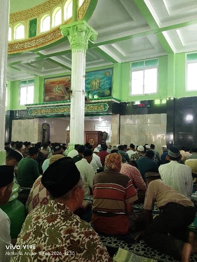 A mosque's interior is filled with men praying. Walls are painted a light green, adorned with decorative paintings above a dark marble section. The ceiling features a light green decorative design, matching the pillars. Natural light filters through windows. The scene captures the atmosphere of a communal prayer service.