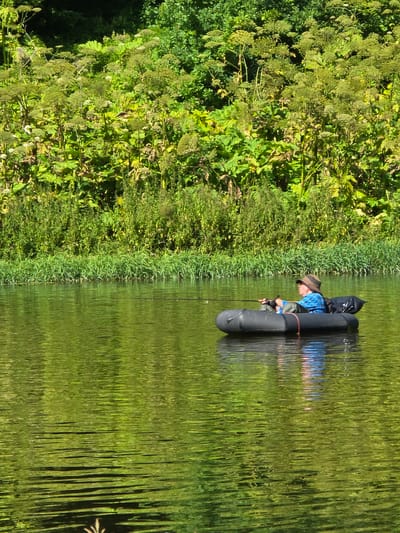 A person wearing a hat and a blue and white checkered shirt is fishing from a black inflatable boat on a body of water. The person is holding a fishing rod. Behind the boat, the bank of the river is covered in lush green vegetation. The water reflects the green of the foliage and the blue of the sky. The scene is lit by sunlight, casting reflections on the water. The image does not contain any specific landmarks or identifiable features that would allow for pinpointing a location within the city of Voinskaya Chast' Mchs, Russia.