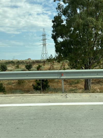This is a vertically oriented, medium-distance, eye-level shot of a roadside scene with a prominent metal guardrail in the foreground, dividing the asphalt road from a grassy, hilly landscape. The road, with a white lane marker, occupies the right side of the frame. To the left of the guardrail, the landscape is dry and overgrown with grass and sparse trees. A large, mature tree with dense green foliage is visible in the upper left quadrant. In the background, a large electricity pylon with visible wires stretches across the sky, which is a pale blue with scattered white clouds. The lighting suggests it is daytime, likely with clear weather. The scene appears to be a rural or semi-rural location, possibly along a highway given the guardrail and pylon. No people are visible. There is a small, red marker on the guardrail. The landscape has a slightly arid appearance, with some structures visible in the far distance on a hillside, including what appears to be a small building with a red roof. The overall impression is one of a journey or passage through a somewhat desolate, yet scenic, environment. The location context provided is Surakhany Raion, Azerbaijan, which aligns with the visual cues of an arid, open landscape with significant electrical infrastructure.