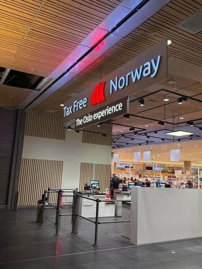 This is a medium-angle, eye-level photograph taken inside an airport terminal, likely at Oslo Airport Gardermoen, Norway, indicated by the "Tax Free Norway" and "The Oslo experience" signage. The scene captures a tax-free retail area with a focus on the entrance to the shopping zone.

The prominent signage, "Tax Free Norway" with a red graphic of what appears to be a stylized sail or wing, is suspended from the ceiling. Below it, "The Oslo experience" is displayed in white lettering. The ceiling itself is constructed of parallel wooden planks with modern lighting fixtures.

In the foreground, there are metal stanchions and barriers, suggesting a queueing or entry system. One of the stanchions has a red laser beam emitting from it, potentially a part of the security or scanning system. To the left, a metal trash receptacle is visible.

Further into the image, a retail store is visible with brightly lit shelves stocked with various products. Several people are present in the background, some appearing to be customers browsing or interacting with staff at checkout counters. The overall environment suggests a commercial, public space within an airport. The lighting is a mix of overhead artificial lights and the glow from the store displays. The floor is dark grey tile.