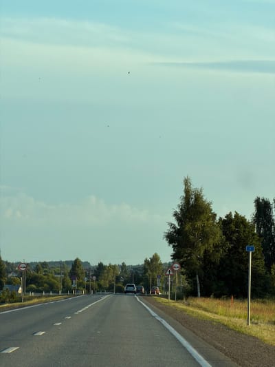 The image shows a two-lane asphalt road in Togashevo, Russia, with white dashed lines marking the lanes and solid white lines bordering the road. The road is lined with trees and vegetation on both sides, and there are several houses visible in the distance on the left. Traffic signs are present along the roadside, including a "No Passing" sign and a "Speed Limit" sign. Several cars are driving on the road, and the sky is a pale blue with some clouds. The overall impression is of a rural road on a clear day.