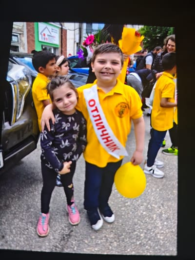 The image depicts a group of children and adults outdoors, likely in a public space. In the foreground, a young boy in a yellow shirt with a white sash that reads "ОТЛИЧНИК" (Excellent) is smiling and holding a yellow ball. Beside him, a young girl in a dark, patterned long-sleeved shirt and black leggings, wearing pink sneakers, is looking at the camera. Another boy in a yellow shirt is standing to the right. In the background, several other people are visible, including adults. There are cars, buildings, and greenery, suggesting an urban or suburban environment. The overall scene appears to be a festive gathering or event. The mention of Kalipetrovo, Bulgaria, in the prompt suggests this location is relevant to the context, although it cannot be visually confirmed from the image alone.