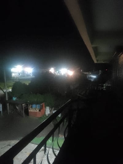 The image displays a nighttime scene in Njoro ward, Kenya, captured from an elevated perspective. In the foreground, a dark metal railing with ornamental scrollwork is visible, extending diagonally from the lower left towards the mid-right. Beneath the railing, a faintly illuminated green surface is present. An overhead, light-colored structural beam or soffit traverses the upper portion of the frame diagonally from left to right.
The midground features a small, partially discernible structure, appearing reddish-brown with a blue section, resting on dark ground that reflects light, suggesting wetness. An indistinct object, possibly a container, is situated to the left of this structure. Dense, dark green foliage occupies the space behind it.
The background is characterized by numerous intense artificial light sources. On the far left, a multi-story building is brightly illuminated by several white lights. To its right, additional clusters of bright white and yellow lights are visible, indicating other structures or activity. The sky is dark, and no individuals or explicit actions are discernible within the frame.