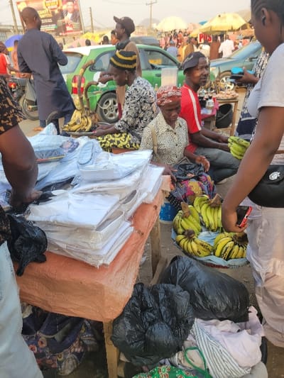 A bustling outdoor market features a variety of goods. A vendor sells bananas alongside packaged white shirts. Black trash bags are piled nearby. Several individuals, including one in a red shirt and another with a yellow-striped hat, are observed amidst the activity. A green vehicle is visible in the background, partially obscured by the market's activity.