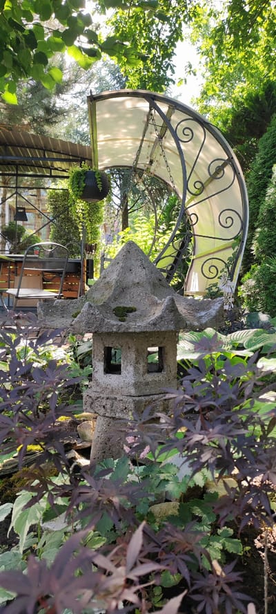 In a garden in Dinskaya, Russia, a concrete pagoda lantern sits amidst lush foliage. The lantern, weathered and covered in moss, features two square openings on its front. Behind the lantern, a white canopy with decorative metal scrollwork is suspended from chains, casting dappled shadows. To the left, a black hanging planter overflows with green vines, and a metal chair and table are visible beneath a corrugated metal roof. The background is filled with various green plants and trees, suggesting a tranquil, verdant setting.
