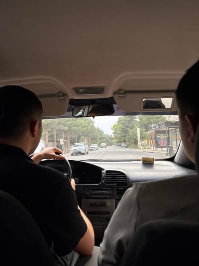 The image, taken from the interior of a vehicle, shows two individuals. The person in the foreground on the left is driving, with their hands on the steering wheel. Their back is to the camera, and they are wearing a black t-shirt. To the right and behind the driver, another person is visible, also with their back to the camera, wearing a grey long-sleeved shirt. The vehicle's dashboard is visible, featuring a car stereo, air vents, and a small yellow ticket on the dashboard. The view through the windshield reveals a street scene. On the right side of the street, there is a bus stop with a sign displaying text and what appears to be the flag of Tajikistan. Trees line the street, and other vehicles are present, including a white car driving ahead and a white sedan further down the road. Buildings are visible in the background. The image appears to have been taken in the city of Guliston, Tajikistan.