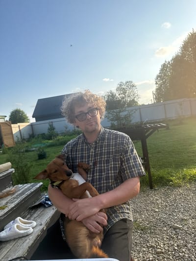 A man with curly hair and glasses, wearing a plaid shirt, holds a small brown dog in his arms. The man is looking at the camera, while the dog is looking away. They are in a backyard with a fence and trees in the background. There are also a pair of slippers on some wooden steps. The sun is setting, casting a warm glow over the scene. The location is the city of Lyady, Russia.