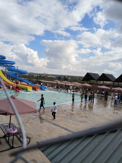The image depicts an active recreational water park situated within Matathani ward, Kenya, under a partially cloudy blue sky.

In the foreground, the upper portion of a gray corrugated metal roof is visible, suggesting an elevated vantage point. A dark gray railing extends horizontally across the lower mid-section. To the left, a large brown patio umbrella shades a wooden picnic table, upon which a small pink inflatable ring rests.

The midground features a large, light-blue swimming pool with visible surface agitation, indicative of a wave pool or active use. Multiple colorful water slides, predominantly blue, yellow, and red, descend into the pool from the left. Several individuals are visible on or at the base of these slides. Numerous people are present in and around the pool area. A shirtless male in dark shorts walks out of the pool onto the wet, light-colored paved deck in the center foreground. Another individual, wearing a white long-sleeved shirt and dark pants, walks away from the pool on the wet pavement. Further back, numerous individuals, including adults and children, are wading in the shallow end of the pool or congregating on the surrounding wet pavement. Brown parasols shade several picnic tables located along the pool's edge and on the paved area. Three larger, dark-roofed, open-sided structures, possibly thatched gazebos, are visible towards the right midground, providing additional shelter.

The background reveals a low wall bordering the water park, beyond which is a sparsely vegetated, seemingly undeveloped landscape. In the far distance, a cluster of buildings forms a skyline against the horizon, indicating a distant urban or semi-urban area.