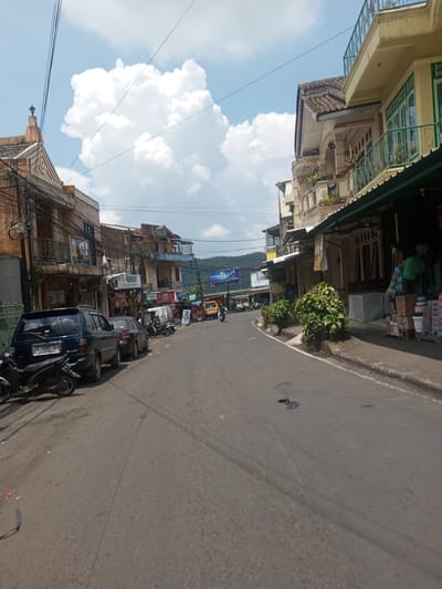 The image depicts a street scene in Xayabury District, Laos. The street is paved and slightly uphill, with parked cars and motorcycles lining the left side. Buildings flank both sides of the street. The buildings on the left are older, with ornate facades and visible utility wires. The buildings on the right appear newer, with balconies and awnings. A person on a motorcycle is visible further up the street. The sky is blue with scattered white clouds.