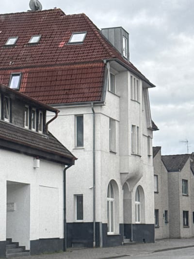 The image captures a street scene in Gronau, Germany, focusing on the architecture of several adjacent buildings. The primary building on the left features a red-tiled, pitched roof with several dormer windows. The facade is predominantly white, with a dark grey base. To the right of this building is a taller, narrower white structure with a more elaborate architectural style, including arched windows and a projecting, curved decorative element. Further to the right, a lighter-colored brick building with a pitched roof and multiple windows is visible, followed by another brick structure. The sky is overcast, and the overall lighting suggests a cloudy day. The foreground shows a paved road and sidewalk. There are no people or vehicles visible in the frame, and no actions or interactions are taking place.