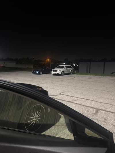The image depicts a parking lot at night in Jeffersontown, United States of America. A dark-colored sedan is parked on the left, and a white SUV is parked to its right. In the foreground, a portion of a vehicle's interior is visible, including a side mirror and window. The window reflects a graphic design resembling a starburst or a spoked wheel. The parking lot is paved with asphalt and shows signs of wear, with visible cracks and markings. Beyond the parking lot, buildings and trees are dimly illuminated by artificial lights. The sky is dark, indicating nighttime.