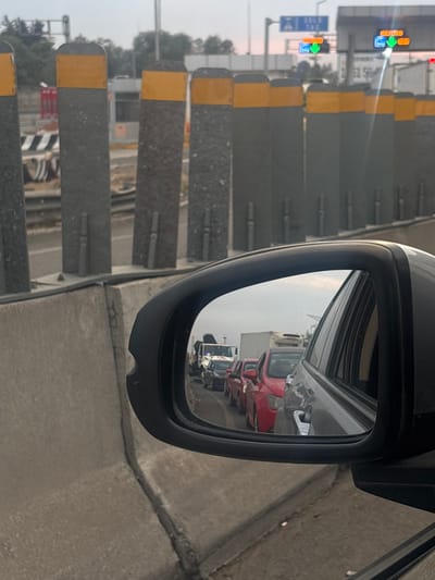 The image depicts a static scene from the interior of a vehicle at a toll plaza on a highway in Tultitlán, Mexico.

The foreground is dominated by the dark-colored exterior side mirror of the vehicle, which occupies the lower right quadrant of the frame. A small chip is visible on the mirror's lower left edge. The mirror reflects a queue of vehicles in a lane behind and to the left of the viewer. This reflected traffic includes a white tow truck with an attached crane, followed by at least three red passenger cars, a dark grey/black passenger car, and a light grey passenger car. Further back, a white box truck or refrigerated truck is visible. The sky in the reflection appears overcast or hazy.

Directly outside the viewer's vehicle, a concrete barrier separates the vehicle's position from the toll plaza infrastructure. Beyond this barrier, a series of tall, grey rectangular pillars with yellow reflective tops are arranged in a line, suggesting a lane divider or part of the toll gate structure. Behind these pillars, components of a toll plaza are visible, including toll booths and an overhead gantry system. Overhead signs on the gantry display "SOLO TAG" in blue, indicating an electronic toll collection lane, along with illuminated green arrows pointing downwards, signifying open lanes. The overall sky is light and hazy. The environment suggests a functional highway setting during daylight hours, characterized by ongoing traffic flow (as seen in the reflection) and toll collection infrastructure. No individuals are explicitly visible in the frame.