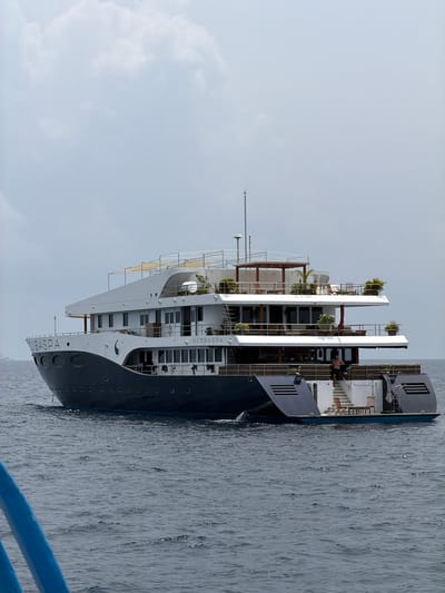 The image shows a ship, likely a cruise ship or large yacht, named "SCOBASPA," on the water. The vessel is multi-decked with some greenery present on the upper levels. It is positioned in the water with a clear sky and some cloud cover in the background. Part of a railing, probably of a smaller vessel from where the picture was taken, is also visible in the lower right corner. The water looks calm with a slightly reflective surface. Based on the presence of vessels and the calm water, the location could be near a harbor or port area, potentially in the vicinity of Maafushi. The image doesn't definitively confirm it is Maafushi, Maldives, but it is plausible based on the environment.