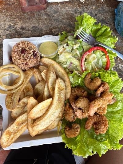A rectangular white plate, presented from a diner's perspective, holds a substantial meal on a textured, weathered brown table in La Ceiba, Honduras. The plate's upper-left section contains a mound of reddish-brown rice mixed with dark red beans. Adjacent to this, a small clear plastic cup contains a pale green, creamy sauce. To the right, a salad composed of torn green lettuce, shredded purple cabbage, and a prominent red bell pepper ring is served, with a silver four-tined fork resting within it. The central and lower-left areas of the plate are filled with numerous elongated, golden-brown slices of fried plantain. On the far left, several golden-fried onion rings are visible. The right side of the plate features a generous serving of breaded and fried shrimp, golden-brown and crispy, arranged on a bed of fresh green lettuce leaves. A blue plastic bag is partially visible in the upper-right background, and the dark-clothed lap of a person is visible beneath the plate in the foreground.
