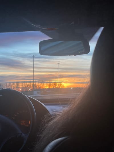 The image captures a vibrant sunset or sunrise viewed from inside a car, likely on a highway near Aubange, Belgium. The perspective looks forward through the windshield, revealing a dramatic sky and road ahead.
In the immediate foreground on the right, a portion of a steering wheel and the dashboard's instrument cluster are visible, though any text or numbers on the gauges are not clearly legible. Human hair partially frames the right side of the view, suggesting a person is either driving or occupying the passenger seat. Centered on the upper left, a rearview mirror reflects a distorted view of the sky or car interior.
Beyond the car, the scene is dominated by a breathtaking sky, awash in brilliant shades of orange and yellow near the horizon, fading into soft purples and blues higher up. This strong coloration indicates either dawn or dusk. A straight road or highway extends into the distance, bordered by trees and sparse vegetation, which appear largely silhouetted against the bright sky. Two tall, dark street light poles stand prominently along the side of the road in the mid-ground. A faint, light-colored structure or building is also visible further down the road on the right. The overall atmosphere conveys a sense of travel during a striking natural light event.