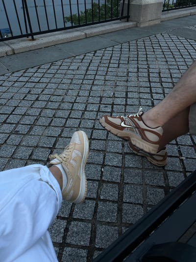 Two individuals are seated on a bench on a cobblestone walkway in Weehawken, New Jersey, with the Hudson River visible in the background behind a black railing. The person on the left wears white pants and tan and cream sneakers. The person on the right has their legs crossed, wearing shorts and brown and tan athletic shoes.