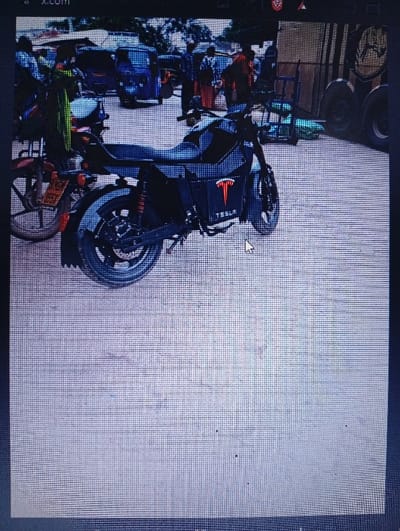 The image is a low-resolution photograph of a digital display, evidenced by screen pixelation and a visible mouse cursor in the lower right foreground. A browser tab displaying "X.com" is partially visible at the top. The scene depicts a street in Shella ward, Kenya.
In the mid-ground, a black motorcycle is prominently positioned, angled slightly to the right, featuring a red "TESLA" logo and text on its side casing. The motorcycle has visible red rear suspension components. To the immediate left of the Tesla motorcycle, the rear section of another motorcycle, including a license plate frame, is partially visible.
The background contains multiple vehicles, including several auto-rickshaws (tuk-tuks), one of which is distinctly blue. On the far right, parts of larger vehicles, possibly a truck or heavy equipment, are discernible. Numerous indistinct human figures are present in the background, appearing as pedestrians or occupants of vehicles. The ground surface is consistent with a paved or unpaved road in an urban or semi-urban environment. No specific actions or interactions are clearly discernible due to image quality.