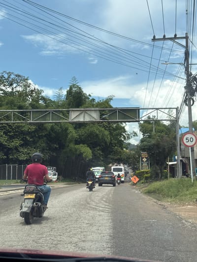 The image presents a daytime scene on a paved road in Pitalito, Colombia, viewed from the interior of a vehicle. The road surface exhibits cracks and wear.

In the immediate foreground, a blurred reddish-brown surface, consistent with a vehicle's dashboard, is visible.

In the midground, a male motorcyclist, wearing a red short-sleeved shirt, blue jeans, and a black helmet, is positioned on the left side of the lane, riding a silver and black scooter-style motorcycle with license plate NXC-97D. Ahead of him, another individual on a dark motorcycle, dressed in dark clothing, proceeds down the road. Behind this second motorcycle, a dark grey SUV (likely a Hyundai Tucson) is in traffic. Further down the road, additional vehicles, including a white van and a white bus, are visible in a line of traffic.

The environment is characterized by dense green vegetation, including tall trees and bamboo, lining both sides of the road and extending into the background. An overhead metal truss structure, appearing to be a pedestrian bridge or sign gantry, spans the road. On the right side of the frame, a utility pole supports multiple overhead electrical wires. A white circular speed limit sign with a red border and the number "50" in red is mounted on a pole to the right. Below this, an orange diamond-shaped warning sign depicting a worker with a shovel indicates road work. A black sign with white text, partially obscured, displays "Delicias Paisas" and business hours, situated to the right amidst the foliage. The sky above is blue with scattered white clouds.

No direct interactions between individuals are discernible. Vehicles are depicted in various stages of forward motion within the traffic lane.