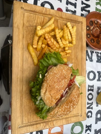 On a light wooden cutting board sits a hamburger cut in half and a pile of french fries. The burger is on the right side of the board, and the fries are on the left. The burger has sesame seeds on the bun and is filled with lettuce, tomato, onion, and an indiscernible sauce. The fries are a medium yellow color.

The background is a patterned tablecloth. Above the cutting board is a brown bowl with black dots arranged in a floral pattern.

The cutting board and food are situated in San Francisco Totimehuacán, Mexico.