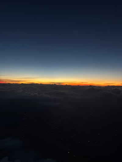 An aerial photograph captures a sunrise or sunset over Santiago Cuautlalpan, Mexico. The upper third of the frame presents a dark blue sky, which transitions to a lighter blue before meeting a prominent horizon band of orange, red, and yellow light. Below this illuminated horizon lies a dense, dark layer of textured clouds, their uppermost edges softly reflecting the vibrant light. The lower half of the image is predominantly dark, revealing scattered, bright points of artificial light on the obscured ground beneath the cloud cover, indicating an urban or semi-urban landscape below at twilight or night. No people, specific objects beyond environmental elements and distant lights, or discernible actions are present.