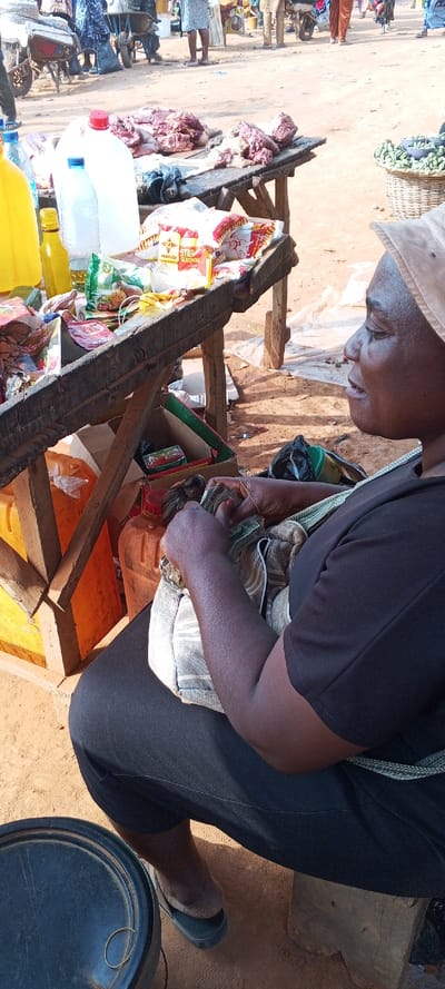 A woman in a black shirt and a sun hat sits on a wooden stool in a busy market. She holds a wad of cash secured with a rubber band in her hands, counting it. Her bag rests on her lap. A wooden table displays various goods for sale, including meat, cooking oil, spices, and other food items. In the background, other people and market stalls contribute to the lively atmosphere.