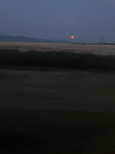 The image shows a twilight sky with a pale blue gradient. A bright red circular object, possibly the moon or sun, is visible in the upper right quadrant of the sky. Below the sky, there is a dark, blurry band that could be a treeline or a dark hill. Further down, a wide expanse of land appears to be a field, with a lighter, textured area in the middle ground and a darker, smoother foreground. In the distance, a faint, dark silhouette of mountains can be seen on the left horizon. Lights from what appear to be buildings are dotted along the horizon on the right side. The overall scene is serene and rural, capturing the quiet moments of dusk or dawn.