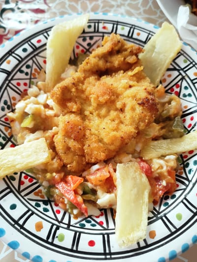 A close-up, overhead shot shows a plate of food. The plate is white with a decorative black, blue, green, and red pattern along the rim. The food consists of a breaded chicken cutlet placed on top of a mixture of chopped tomatoes, peppers, and what appears to be scrambled eggs. Several pieces of fried plantain are scattered around the chicken. The background is blurred but suggests a patterned tablecloth. The image is taken indoors.