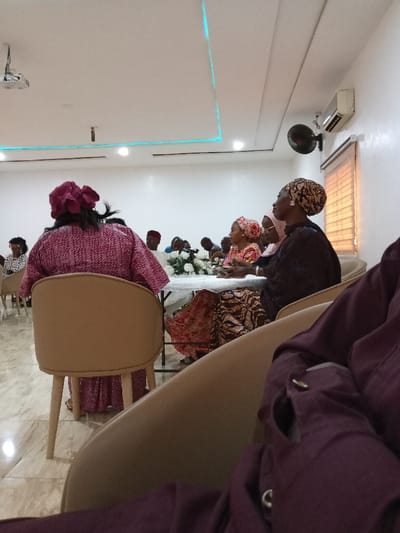 The image shows a group of people seated at a table in a room with white walls and a tiled floor. There are beige chairs and a white table with white flowers on it. The room has a white ceiling with blue lights and there is a ceiling fan with a brown blade in the upper right hand corner of the room. There is a window with a brown blind behind the table.  The people are dressed in traditional clothing. The people are facing the table and talking to each other. There is a woman wearing a red and white dress, another woman wearing a red and black dress, and another woman wearing a blue and white dress. There is a man wearing a white and black dress shirt and a blue tie. The woman in the red and white dress is seated in a beige chair facing the camera. The woman in the red and black dress is seated in a beige chair facing the other people. The woman in the blue and white dress is seated in a beige chair facing the other people. The man is seated in a beige chair facing the other people. The image was taken in Yola, Nigeria, at the coordinates 9.198, 12.491. The image was taken indoors. The image was taken during the day. The image was taken in a well-lit room. The image was taken from a seated position. The image was taken looking forward. The image was taken with a smartphone.