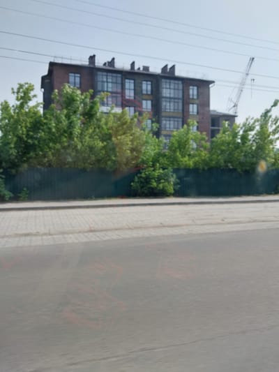 The image depicts a building under construction, likely in Belokurikha, Russia, given the context. A large crane is visible in the background, suggesting ongoing construction work. The building itself appears to be a multi-story structure with a brick facade and large windows. Trees and foliage surround the building, indicating an outdoor setting. The sky is overcast, hinting at potentially cloudy or rainy weather. The image also shows a paved surface, possibly a road or sidewalk, with faint red markings that could be graffiti or chalk lines. No people are clearly visible in the frame, but the presence of construction equipment implies human activity is occurring or has recently occurred. The overall impression is one of an industrial or urban development site. The orientation of the image is rotated 90 degrees counterclockwise.