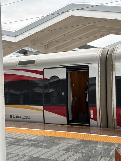 The image shows a train car at a station in Yongding District, China. The train car is painted white, black, and red. The number "ZE 323403" and the words "Second Class Coach" are printed on the side of the train car. The train door is open. A person is inside the car. The station platform is made of gray stone with a yellow tactile warning strip. A building with a geometric architectural style stands next to the train station.