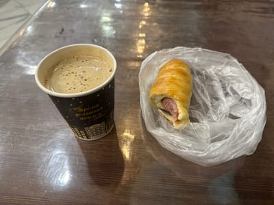 The image shows a close-up, eye-level view of a coffee cup and a hot dog wrapped in a bun, placed on a wooden table. The coffee is in a black disposable cup with a white rim, filled with a frothy, light brown liquid. The hot dog is encased in a golden-brown, spiral-shaped bun, with the pink sausage visible at the open end. It is partially covered by a clear plastic bag. The table surface is dark brown with a visible wood grain and reflects light, suggesting it might be a laminate or polished wood. The background is blurred, indicating a shallow depth of field. The lighting appears to be artificial, casting shadows and highlights on the table. The scene is devoid of any human presence or activity. The image does not provide any specific landmarks or contextual clues that would associate it with the city of Inzer, Russia, or any other specific location.
