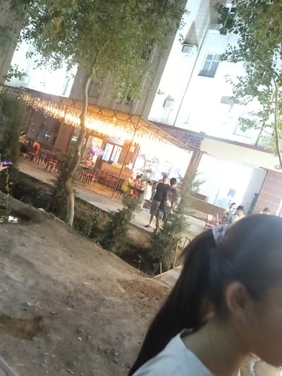 The image appears to depict an outdoor seating area of a cafe or restaurant in Kungrad, Uzbekistan. The scene is illuminated by string lights overhead, creating a warm ambiance. Several tables and chairs are arranged on a paved area, some of which are occupied by patrons. In the background, a multi-story building with windows and air conditioning units is visible. There are trees and other foliage scattered throughout the scene. In the foreground, the back of a woman's head is visible, with her hair tied in a ponytail. The ground in the foreground appears to be dirt or gravel, with some scattered debris. The overall impression is that of a lively evening gathering at a local establishment.