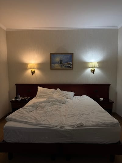 This image depicts a dimly lit hotel room in Zhravlevo, Kemerovo, Russia. The primary subject is a large, unmade bed with rumpled white linens that occupies a significant portion of the frame. The bed is positioned against a headboard that appears to be made of dark wood. To the left of the bed, on a wall with textured wallpaper, are two wall-mounted lamps. The upper lamp casts a warm glow on the wall, while the lower lamp illuminates a framed painting of a harbor scene with a windmill. The room also features a small bedside table with a water bottle and what appear to be other assorted items. The overall impression is one of a room that has recently been occupied, possibly late at night or early in the morning, given the low light levels and the unmade bed. The scene is static, with no discernible activity or people present. The time of day is suggested to be evening or night due to the artificial lighting.