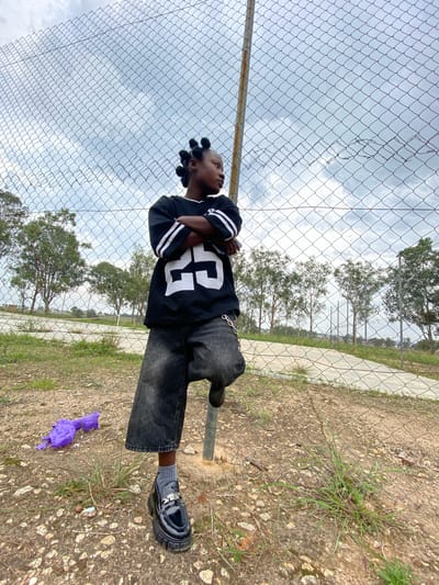 A young person with braided hair stands with their arms crossed next to a chain-link fence in Zawan, Nigeria. They are wearing a black jersey with white stripes on the sleeves and the number "25" in white on the front, along with wide-leg dark denim pants and black platform loafers. A silver chain is attached to the left side of their pants. A crumpled purple plastic bag is on the ground to the left of the person. The background shows a cloudy sky, trees, and a paved pathway.