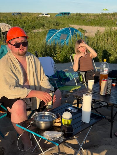 The image captures a sunny outdoor scene on a sandy beach, likely in Povarovka, Russia, given the context. In the foreground, a man wearing an orange hard hat and sunglasses is seated in a lawn chair. He is draped in a light-colored towel and holding a can of soda. To his right, a black folding table holds several items, including a metal bowl, bread, a roll of paper towels, a small light, a pack of cigarettes, and a portable speaker. Further back, a young woman with blonde hair is seated in a green lawn chair, with her hand shielding her eyes from the sun. Next to her, a large plastic bottle of amber liquid and two cans are visible on a table. In the background, a blue tent is set up, along with other camping gear and what appear to be parked cars and a green umbrella. The environment is characterized by tall grasses and a sandy beach under a clear sky with warm, late afternoon sunlight casting long shadows.